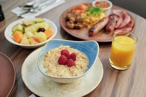 Ein Frühstückstisch mit einer Schüssel Müsli, belegt mit Himbeeren, einem Glas Orangensaft sowie Tellern mit Speisen, darunter Würstchen und Obstsalat.