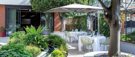 An outdoor dining area with tables and chairs, surrounded by greenery and a tree.