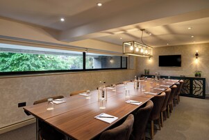 A conference room with a long table, chairs, a TV, and a window with a view of greenery.
