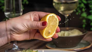 A hand holding a lemon slice over a plate of food, with a glass of white wine and a bowl in the background.