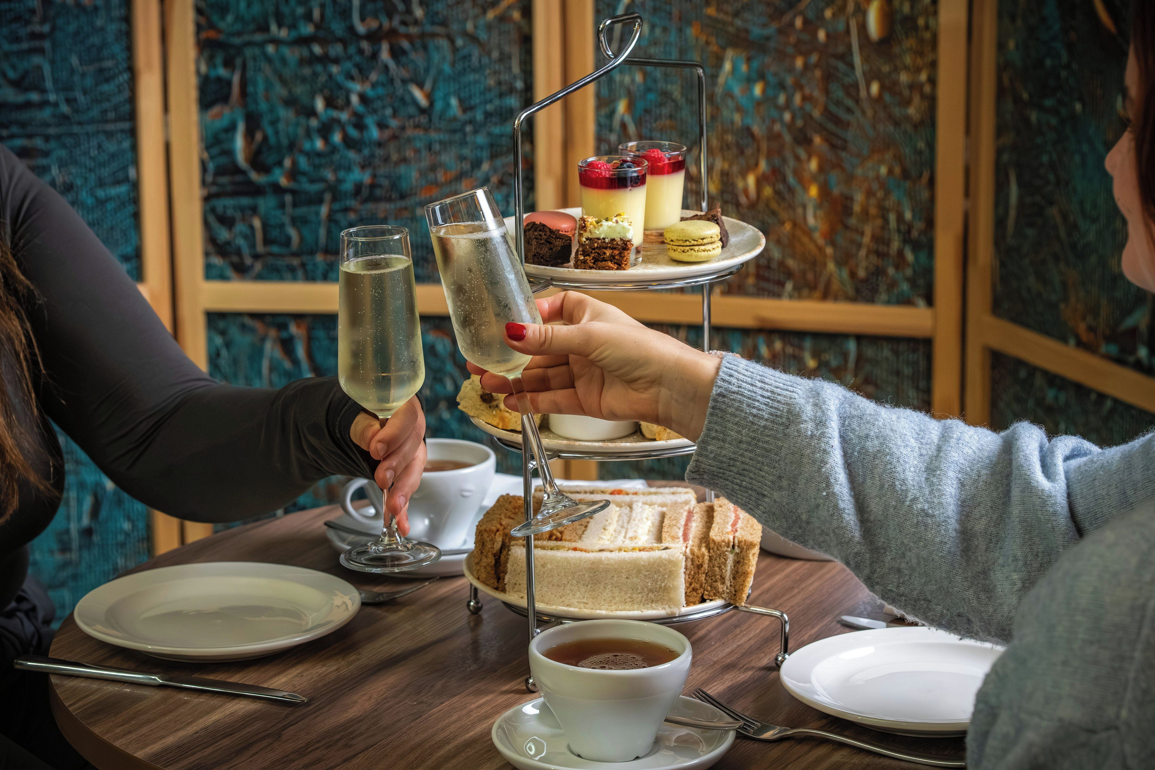 Una mesa con un soporte escalonado para postres, dos vasos de una bebida transparente, una taza de té y un sándwich.