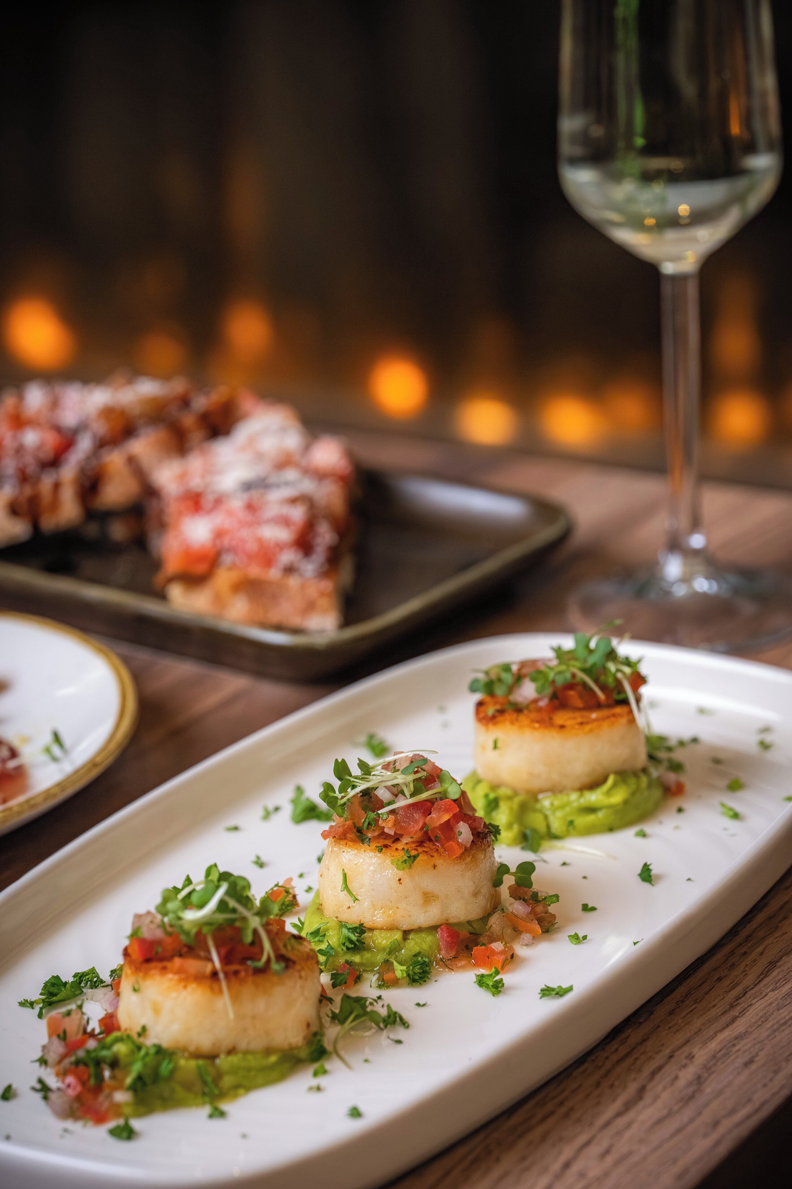 Um prato retangular branco com três vieiras decoradas com ervas e vegetais, um copo de vinho branco, e um fundo desfocado com uma mesa de madeira.