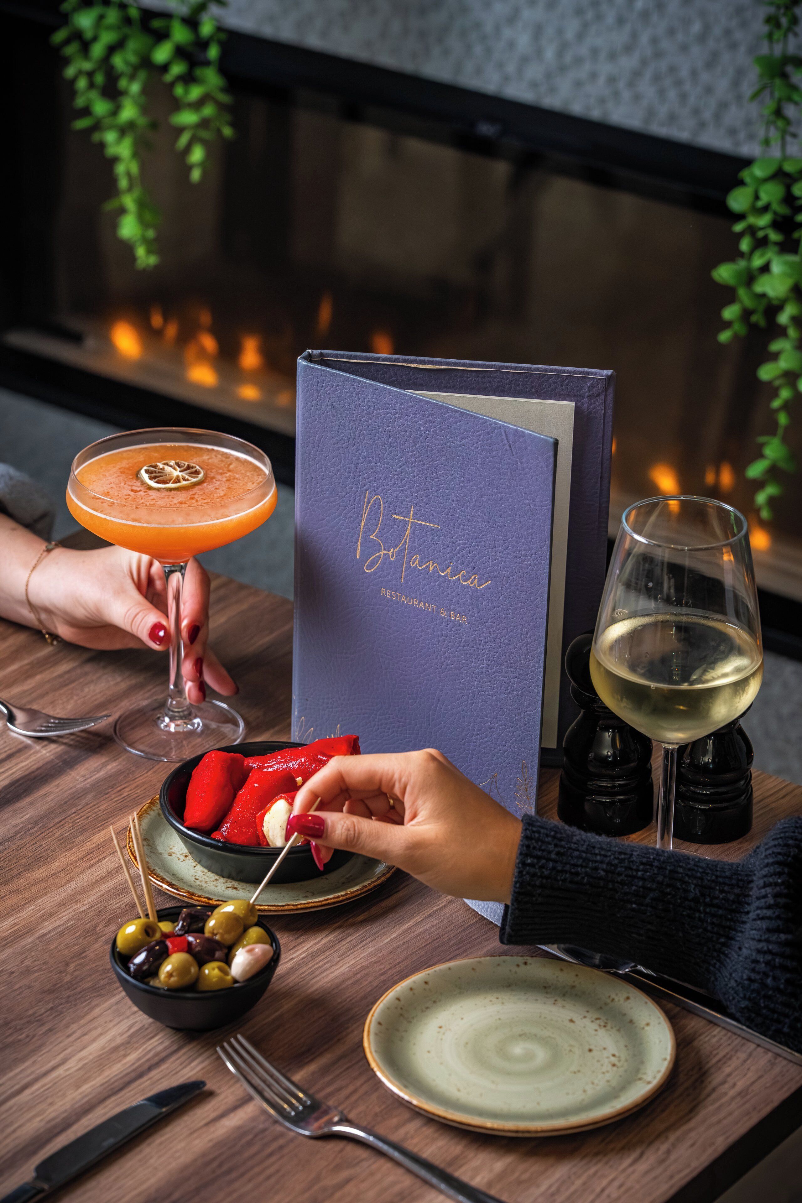 A table with a menu, a cocktail, a glass of white wine, a bowl of olives, and a plate with strawberries.