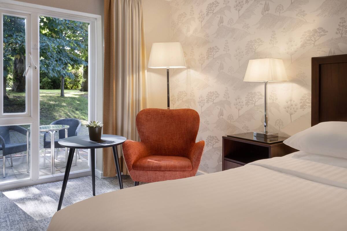 Une chambre d’hôtel avec un lit, deux lampes, une chaise, une petite table avec une plante et une vue sur l’extérieur par une fenêtre.