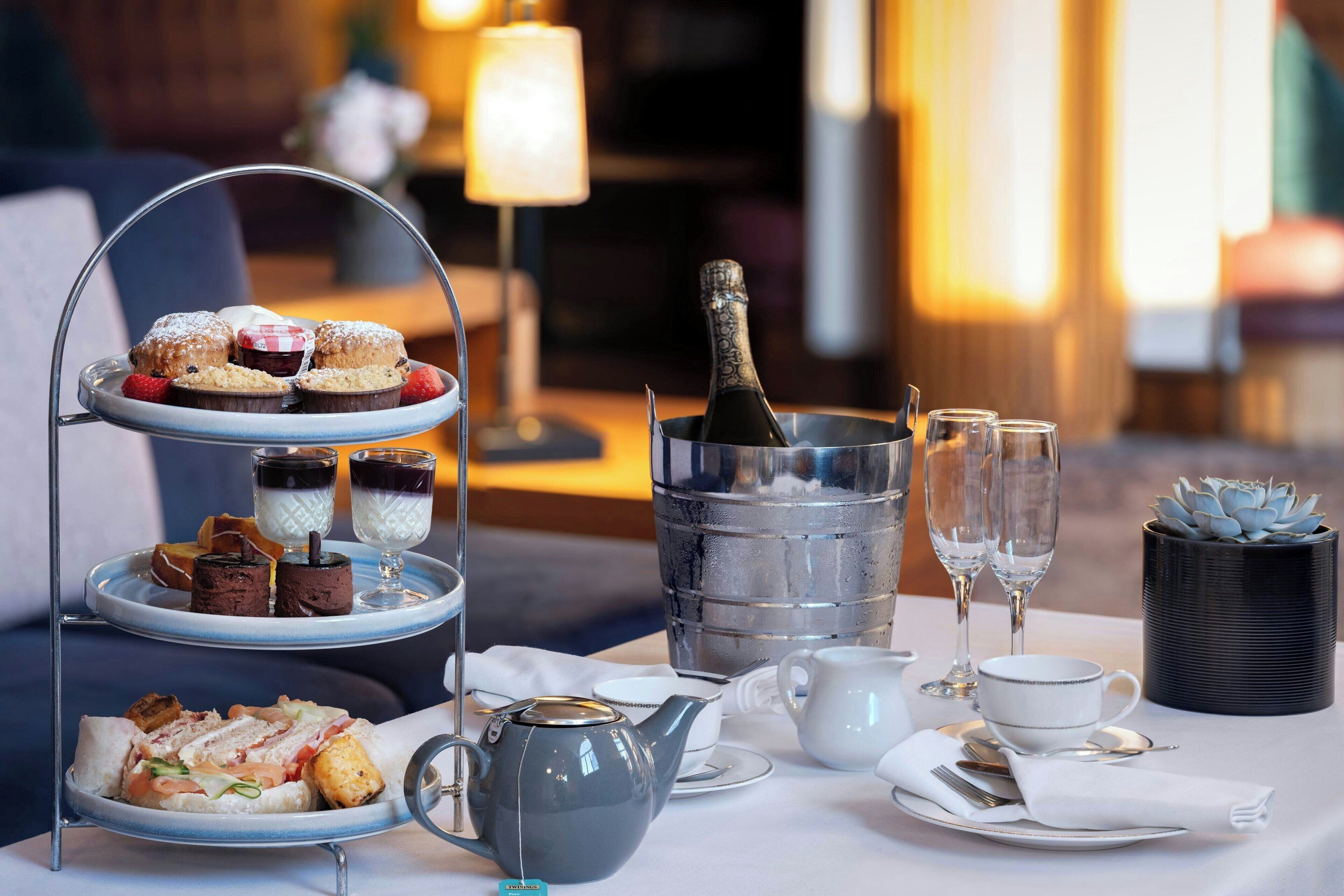 A three-tiered dessert stand with various pastries, a bottle of champagne, and two glasses of champagne on a table set for a meal.