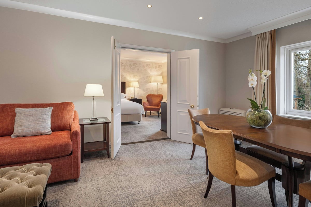 Une chambre d’hôtel équipée d’un canapé, d’une table à manger, d’une chaise, d’une lampe, d’un lit et d’une fenêtre avec des rideaux.