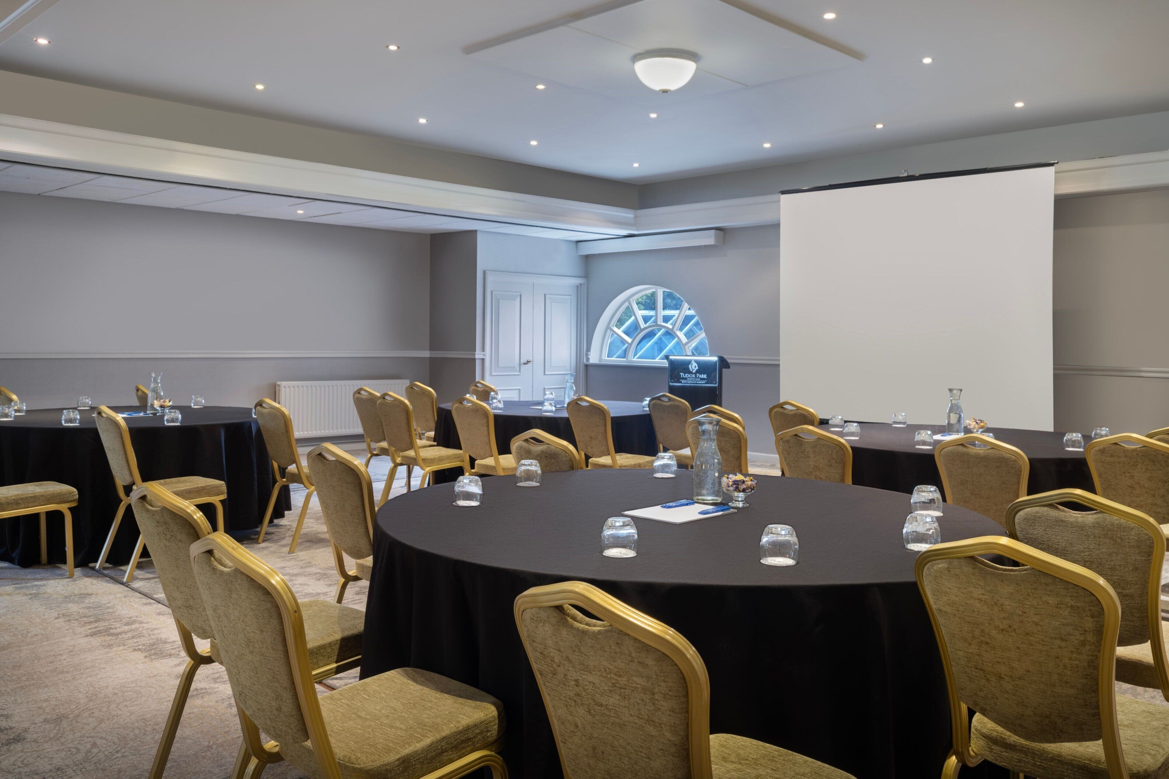 Un salón de conferencias con mesas redondas, sillas y una pantalla para proyector.