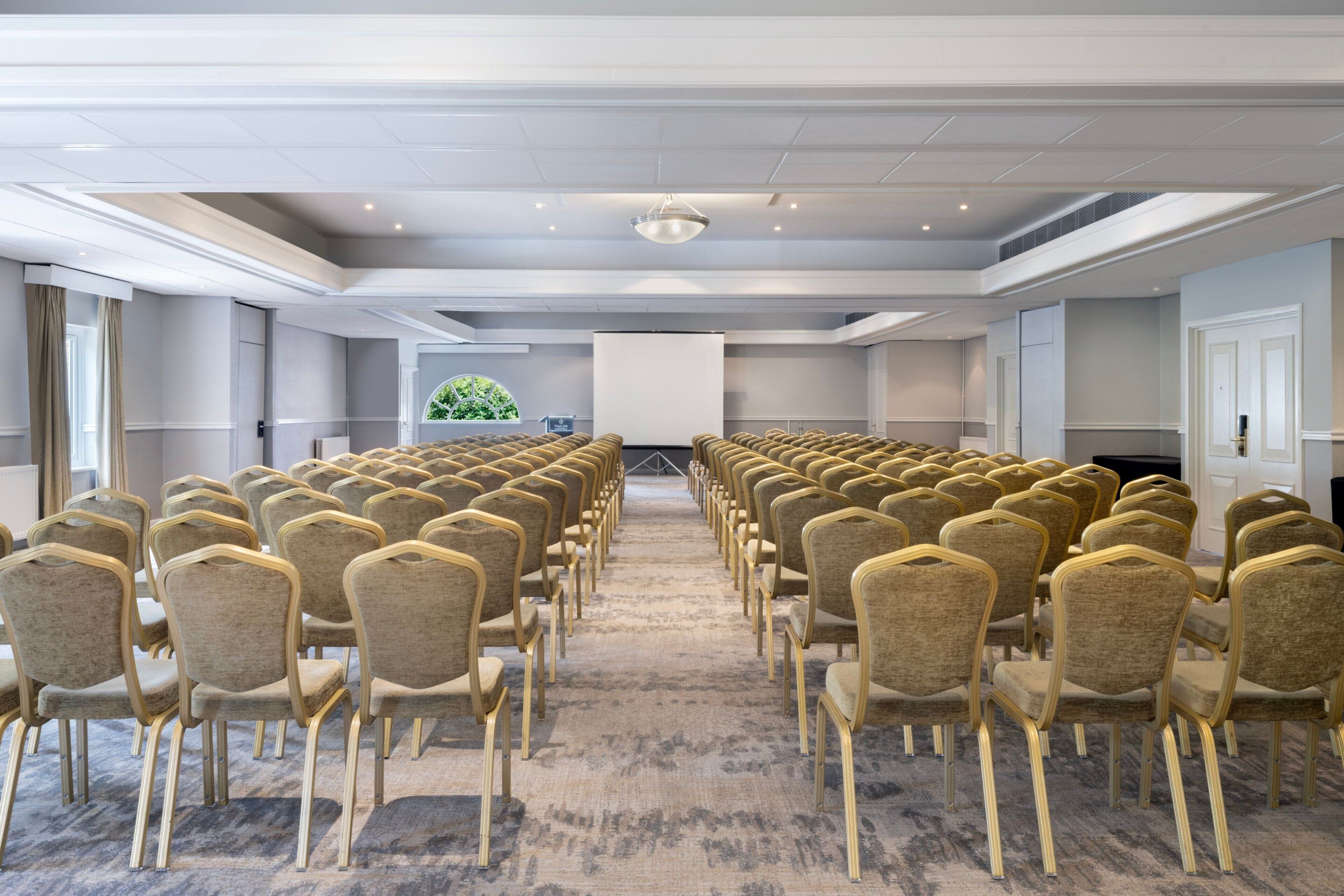 Une salle de conférence avec des rangées de chaises disposées face à un grand écran, un luminaire au plafond et une vue sur un paysage verdoyant par la fenêtre.