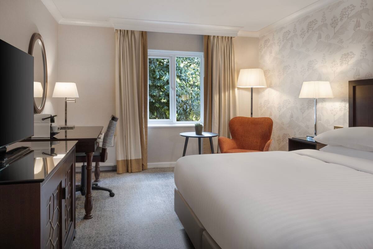 Une chambre d’hôtel avec un grand lit, un bureau avec une télévision, deux lampes, une chaise et une fenêtre avec des rideaux.