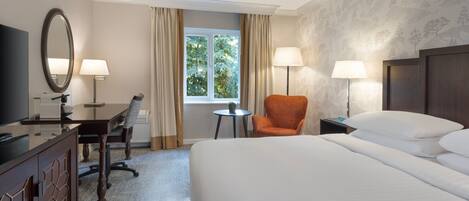 A hotel room with a bed, a desk with a chair, a TV, two lamps, and a window with curtains.