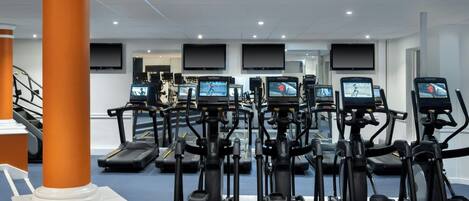 A gym with multiple treadmills and elliptical machines, a staircase, and a flat-screen TV mounted on the wall.