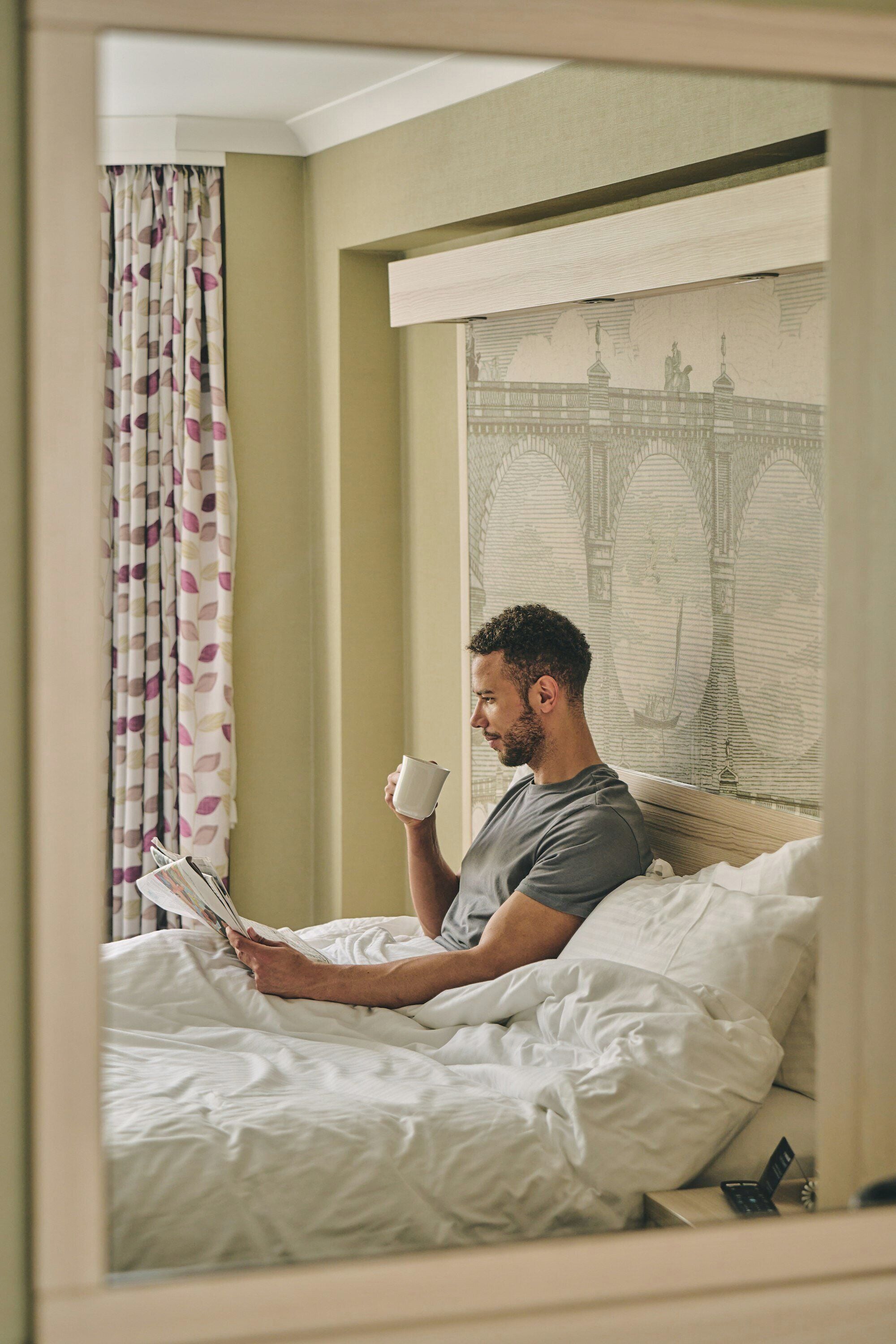 Iemand leest een boek in bed, met een kopje in de hand. Er is een raam met gordijnen en een muur met een muurschildering van een brug.