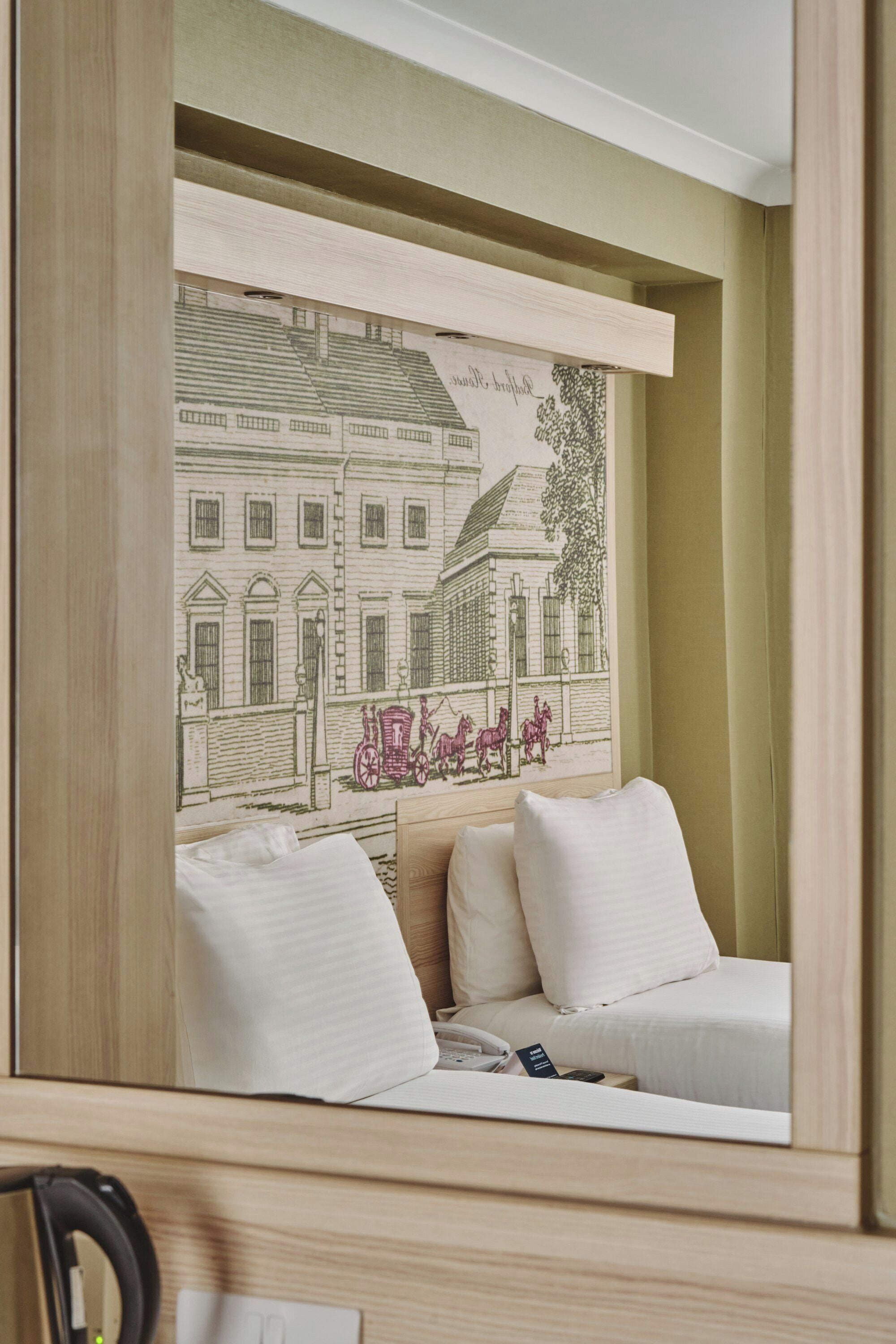 A hotel room with a bed, a bedside table, and a wall-mounted artwork depicting a building and horses.