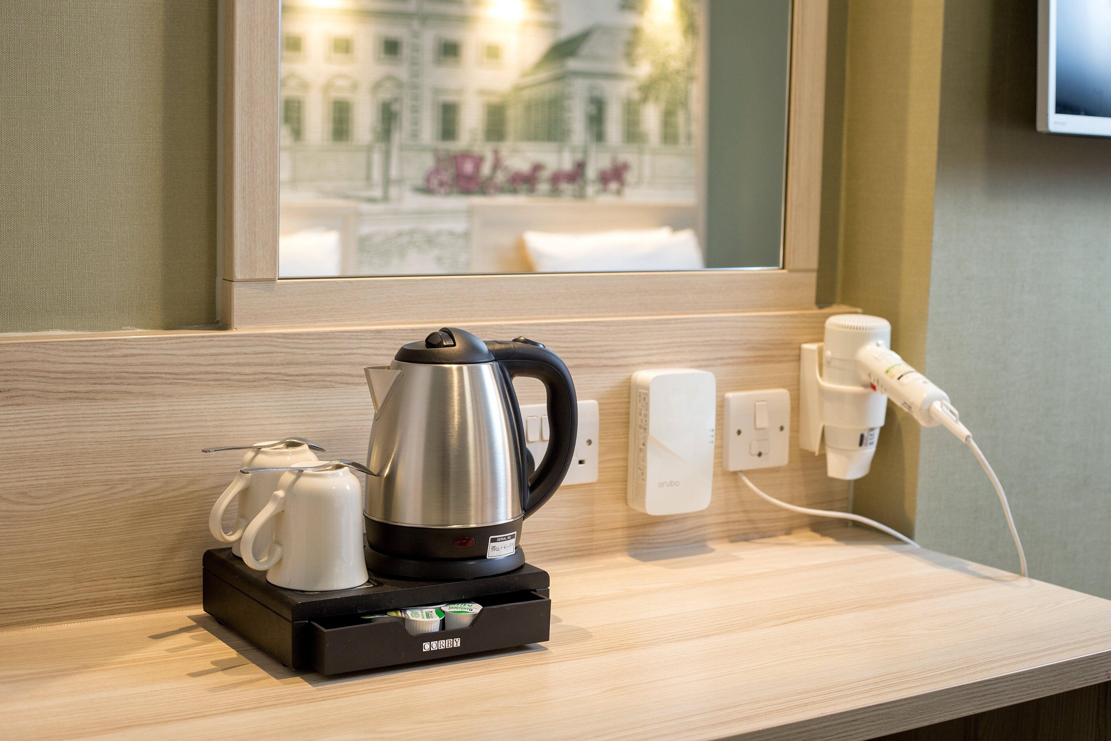 A coffee maker with a kettle and a cup on a wooden counter, a power outlet, and a wall-mounted device.