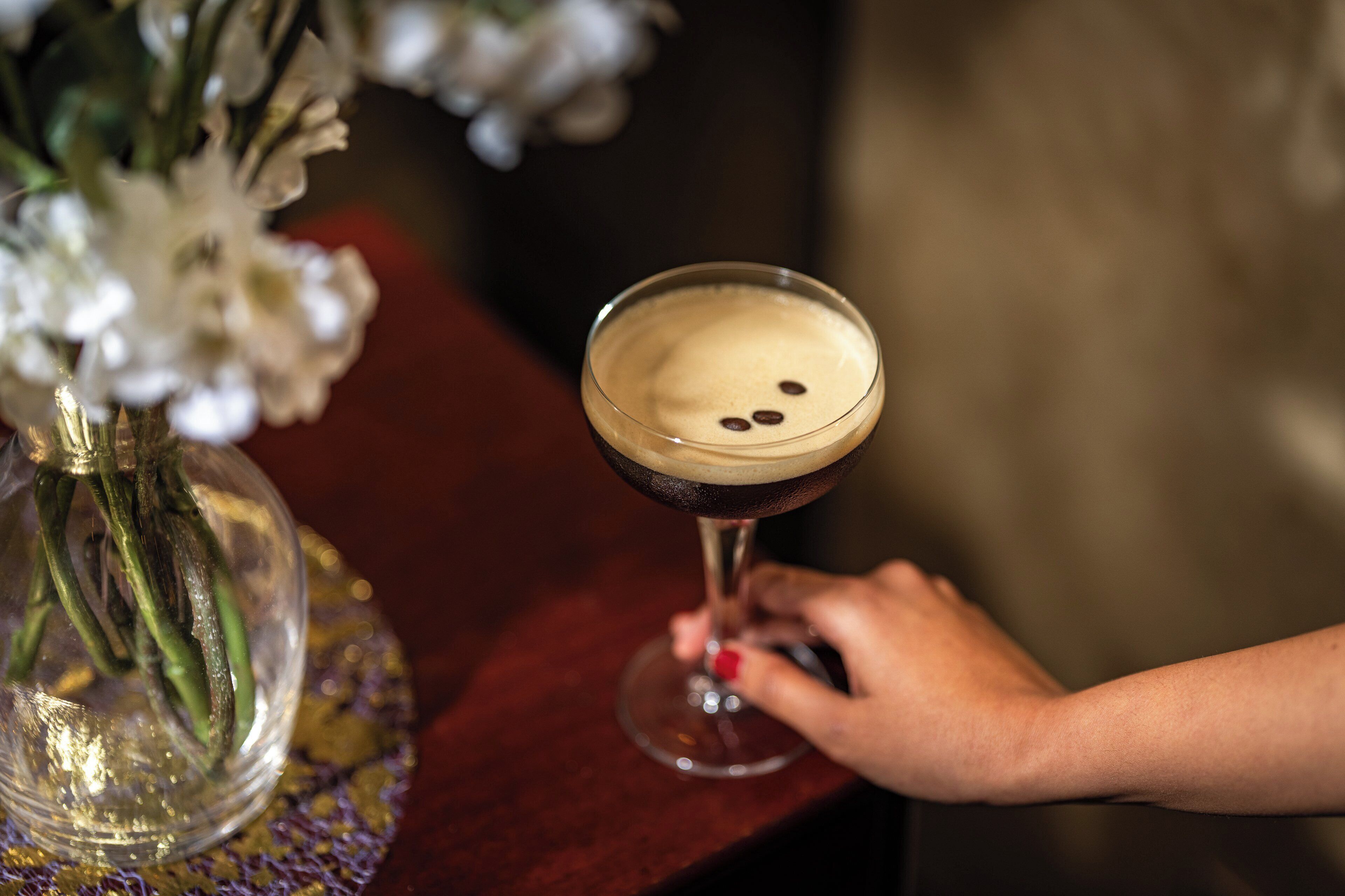 Käsi, jossa on cocktail-lasi, jossa on tummaa nestettä ja vaahtoa, ja lasi on asetettu puupinnalle.
