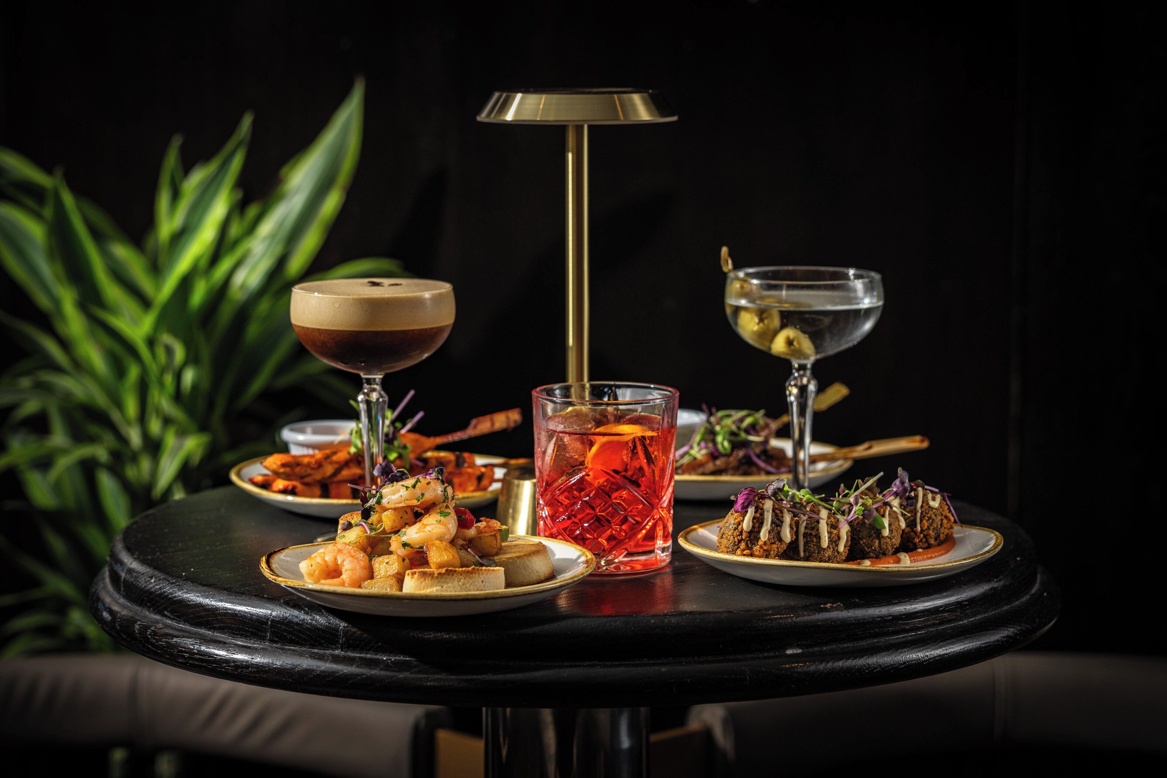 A round table with plates of food, glasses of beverages, and a tall lamp.
