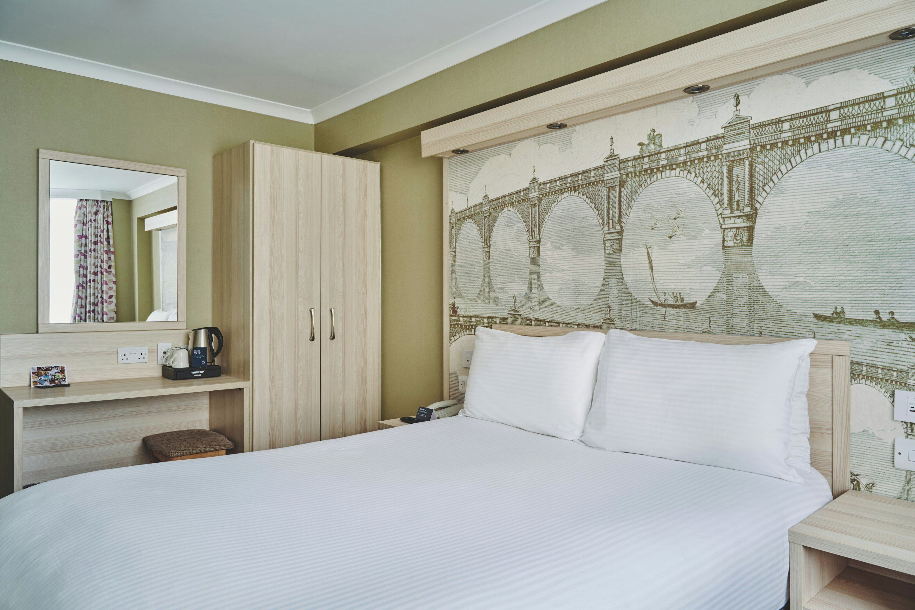 A hotel room with a bed, a wooden wardrobe, a bedside table with a coffee maker, and a large mural of a bridge.