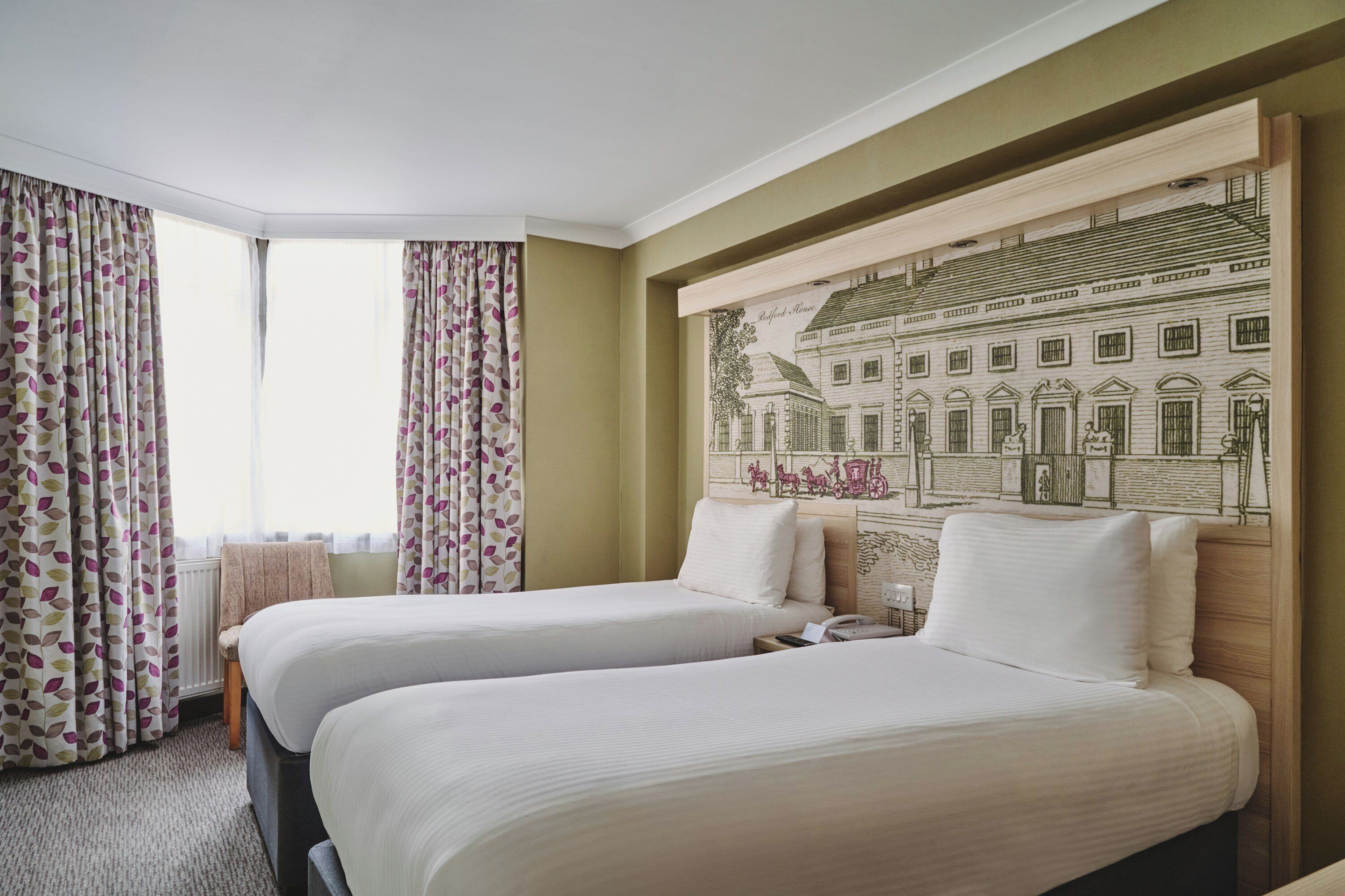 Ein Hotelzimmer mit zwei Betten, einem großen Fenster mit Vorhängen und einem Wandbild eines Gebäudes.