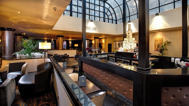 A spacious lobby with a high ceiling, a bar area, seating arrangements, and a large window providing natural light. - President Hotel (London)