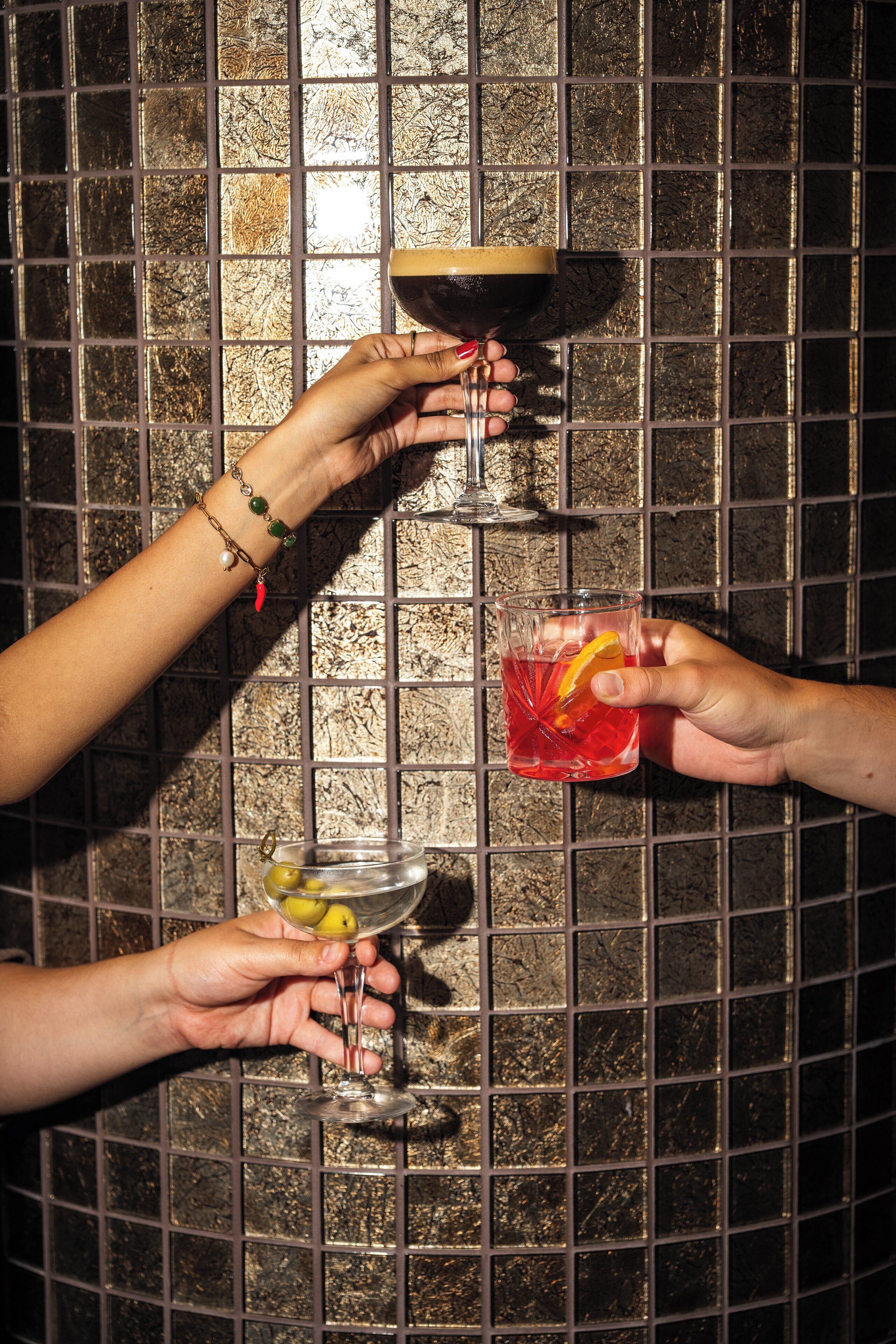 Tres manos sostienen diferentes vasos de bebidas frente a una pared de azulejos.