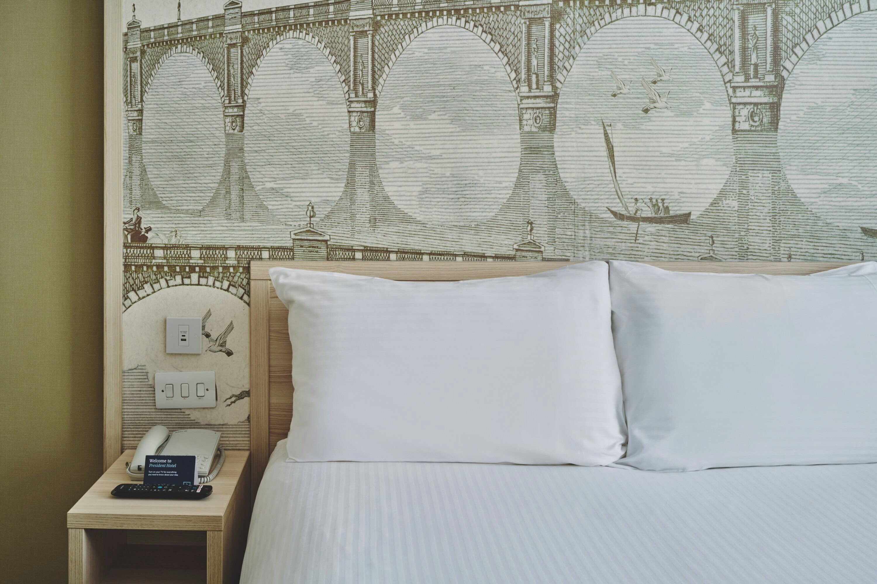 Una camera d'albergo con un letto, cuscini, un comodino con un telefono, un cartello di benvenuto e un murale raffigurante un ponte.
