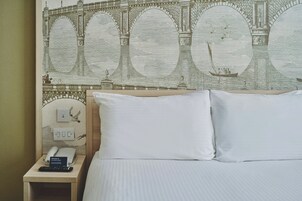 Ein Hotelzimmer mit Bett, Kissen, einem Nachttisch mit Telefon, einem Willkommensschild und einem Wandbild einer Brücke.