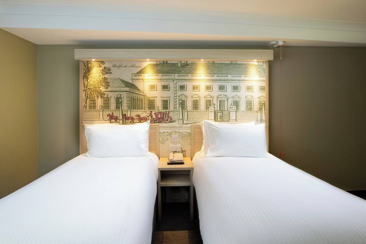 A hotel room with two single beds, a nightstand with a lamp, and a wall mural depicting a street scene.