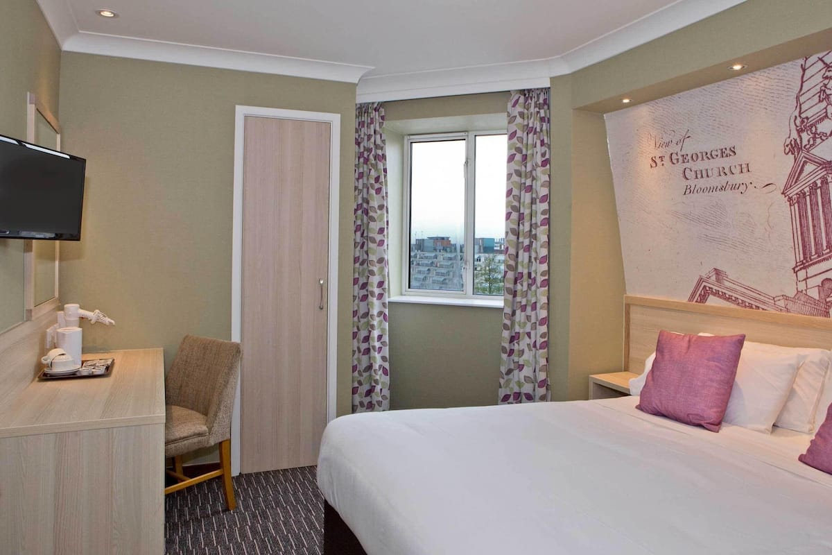 A hotel room with a bed, a desk with a chair, a television, a window with curtains, and a wall with a printed image.