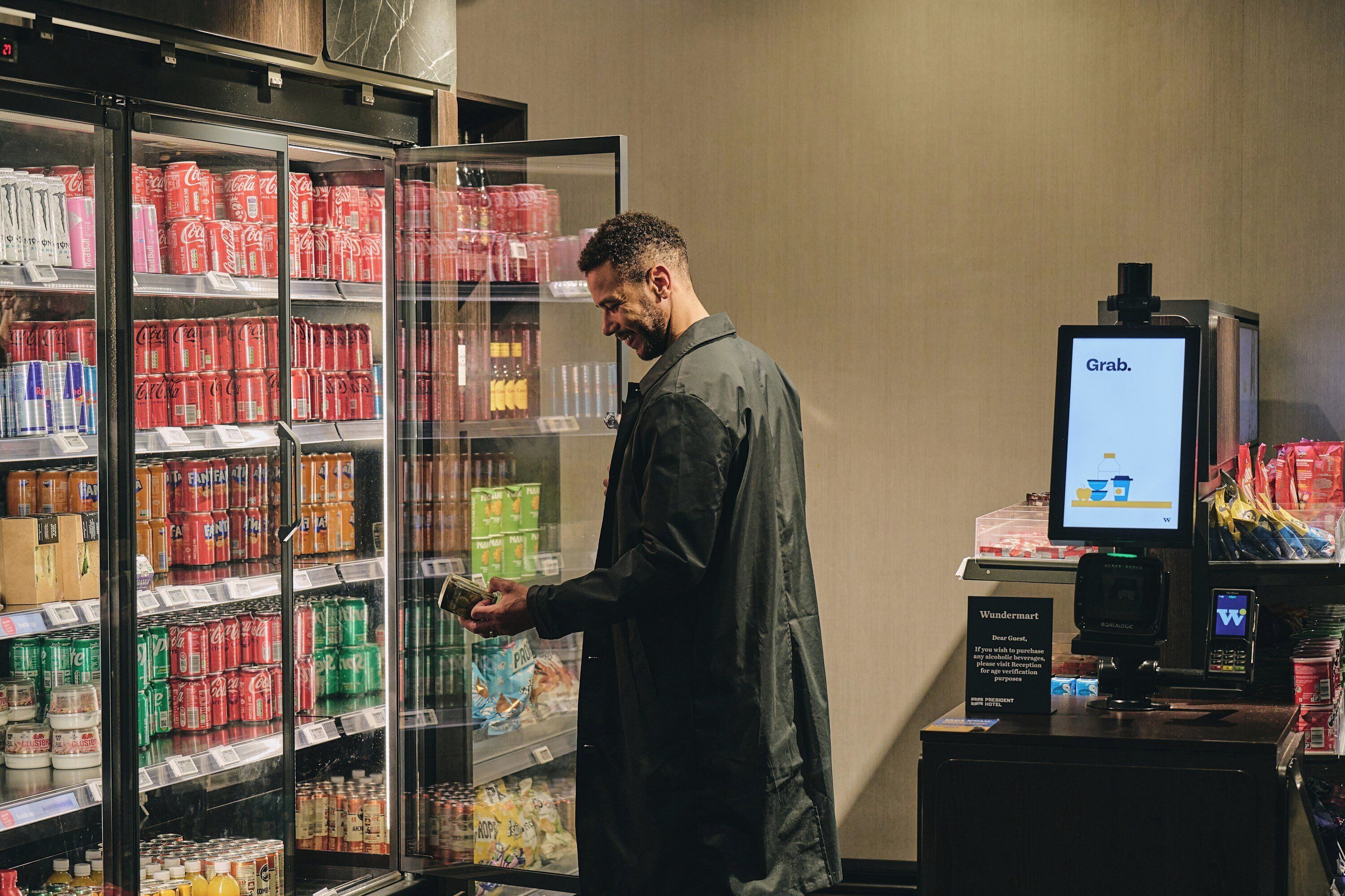 A person in a dark jacket standing in front of a refrigerated beverage display with various drinks and a digital screen displaying 'Grab.'