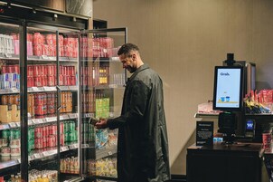 A person in a dark jacket standing in front of a refrigerated beverage display with various drinks and a digital screen displaying 'Grab.'
