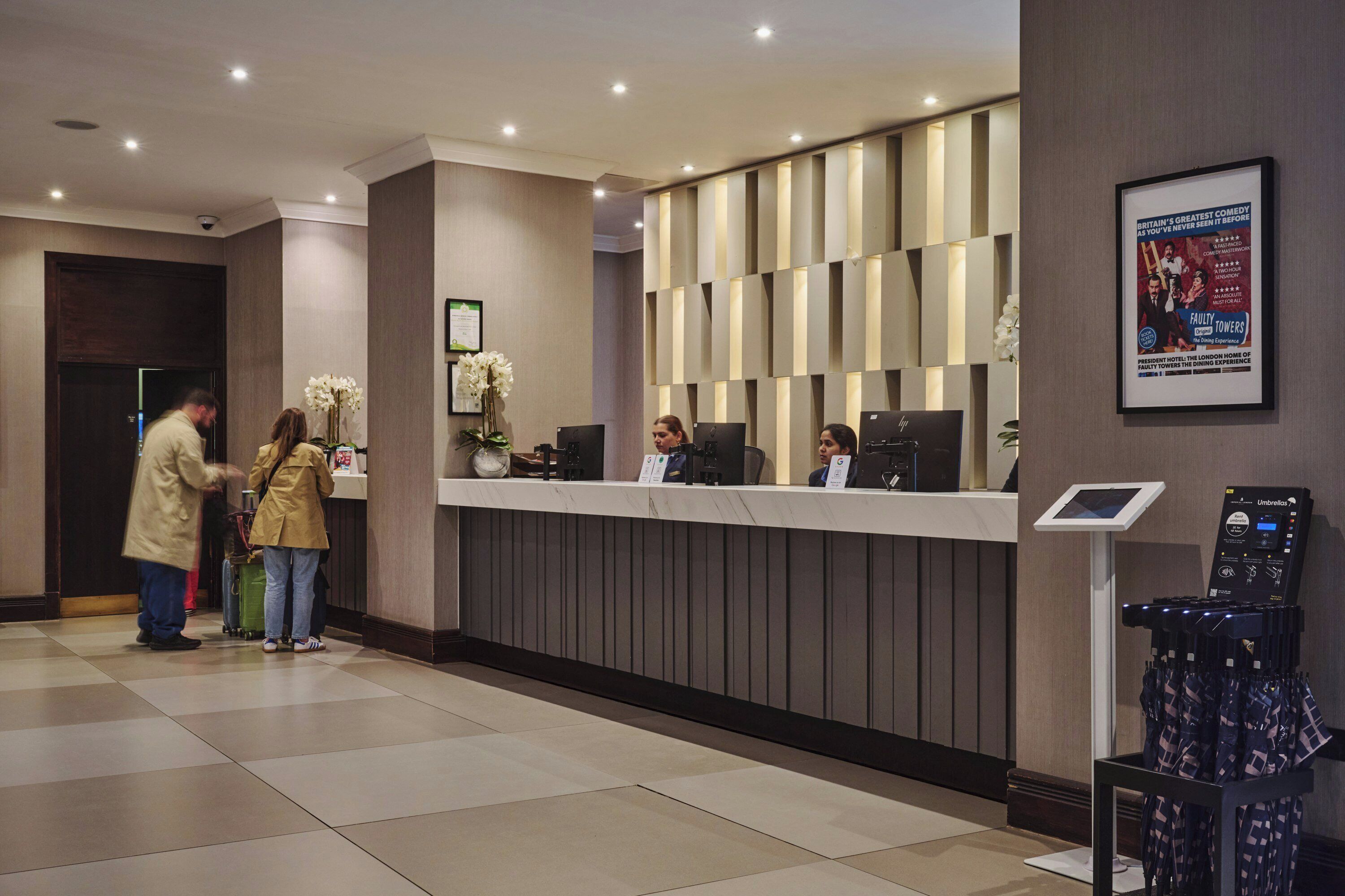 Zona de recepción de un hotel con mostrador de entrada, quiosco digital y un cartel enmarcado en la pared.