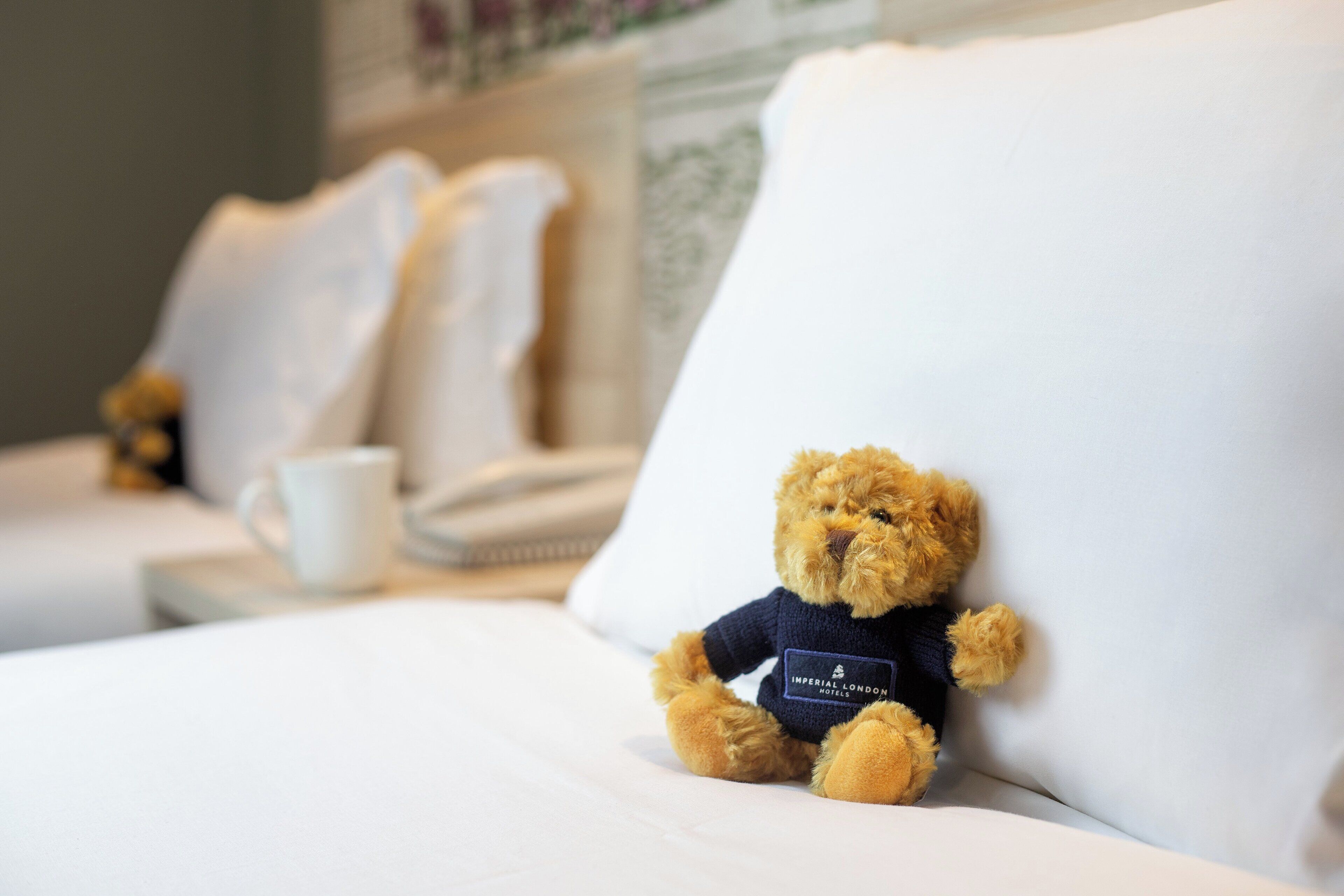 A plush toy on a bed with pillows and a cup in the background.