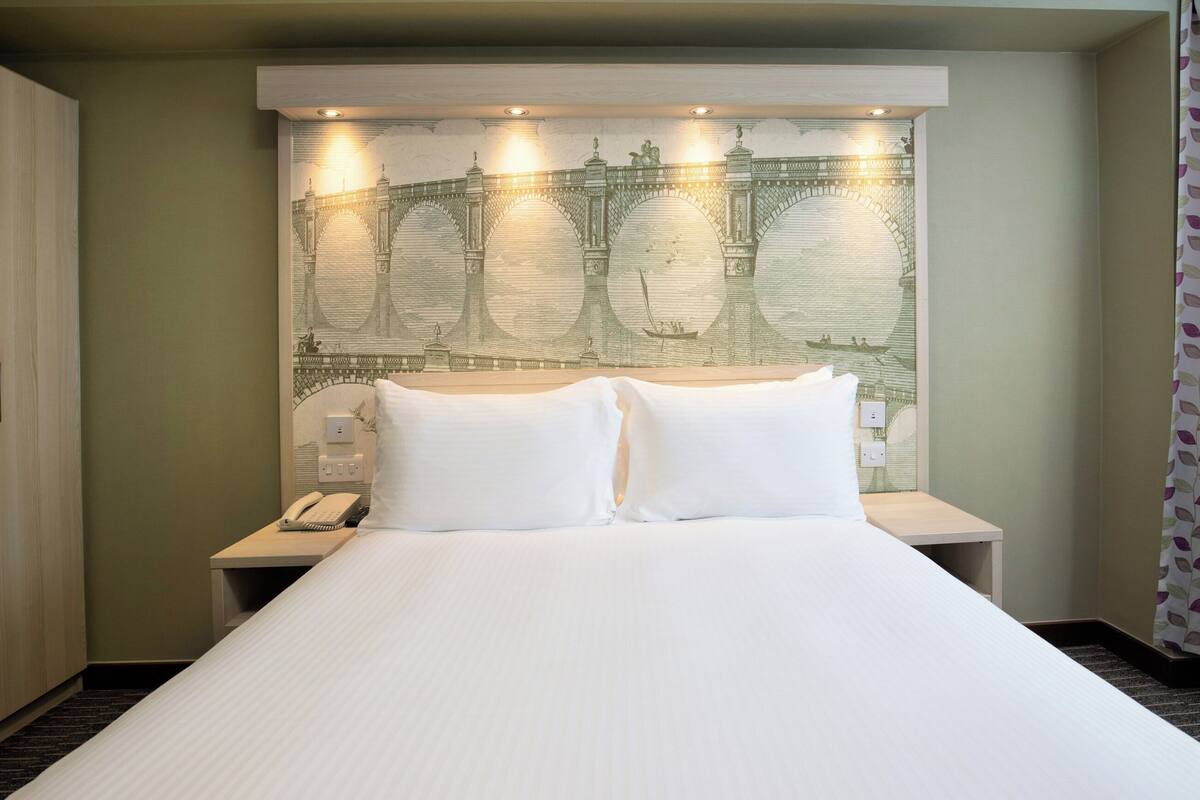 A hotel room with a large bed, a headboard with a mural, a bedside table with a phone, and a curtain with a leaf pattern.