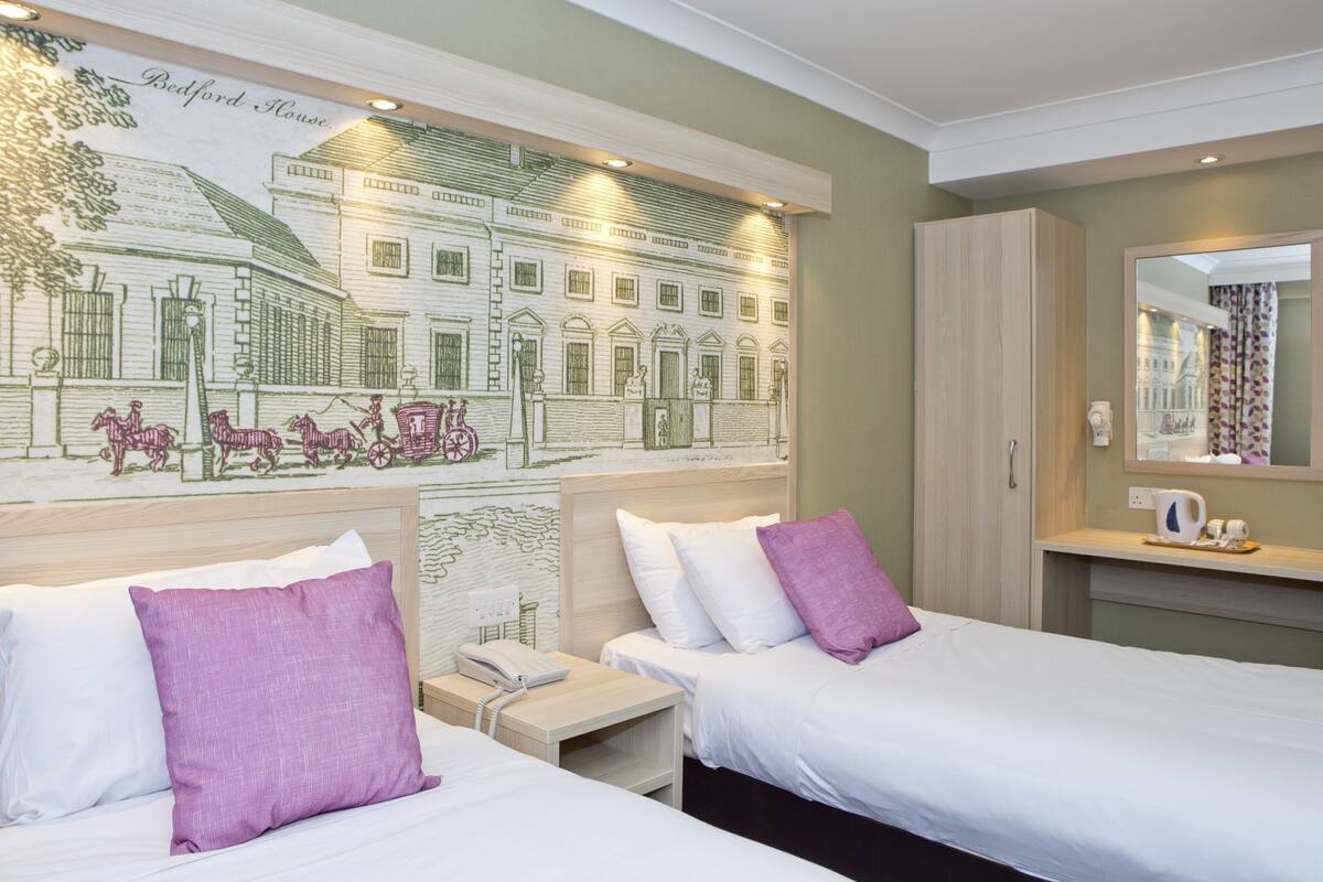 A hotel room with two beds, a headboard with a mural, a bedside table with a phone, and a mirror with a curtain.
