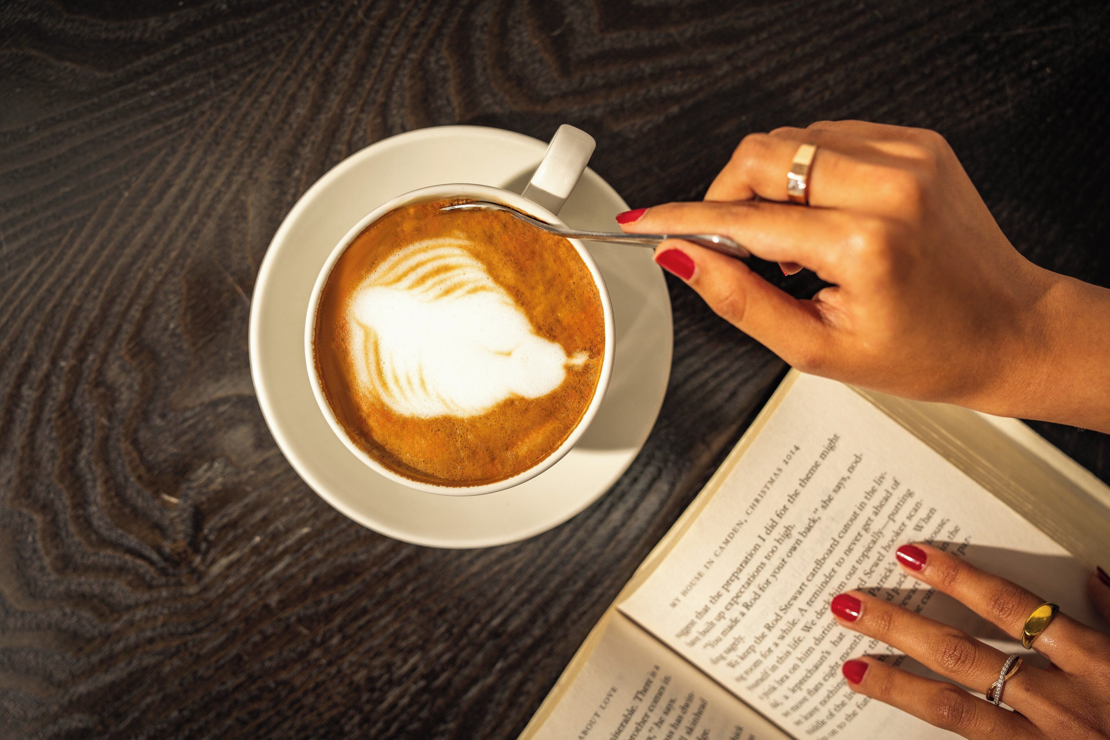 Une tasse de café ornée d’un motif en latte art, posée sur une soucoupe, un livre et une main portant une bague.