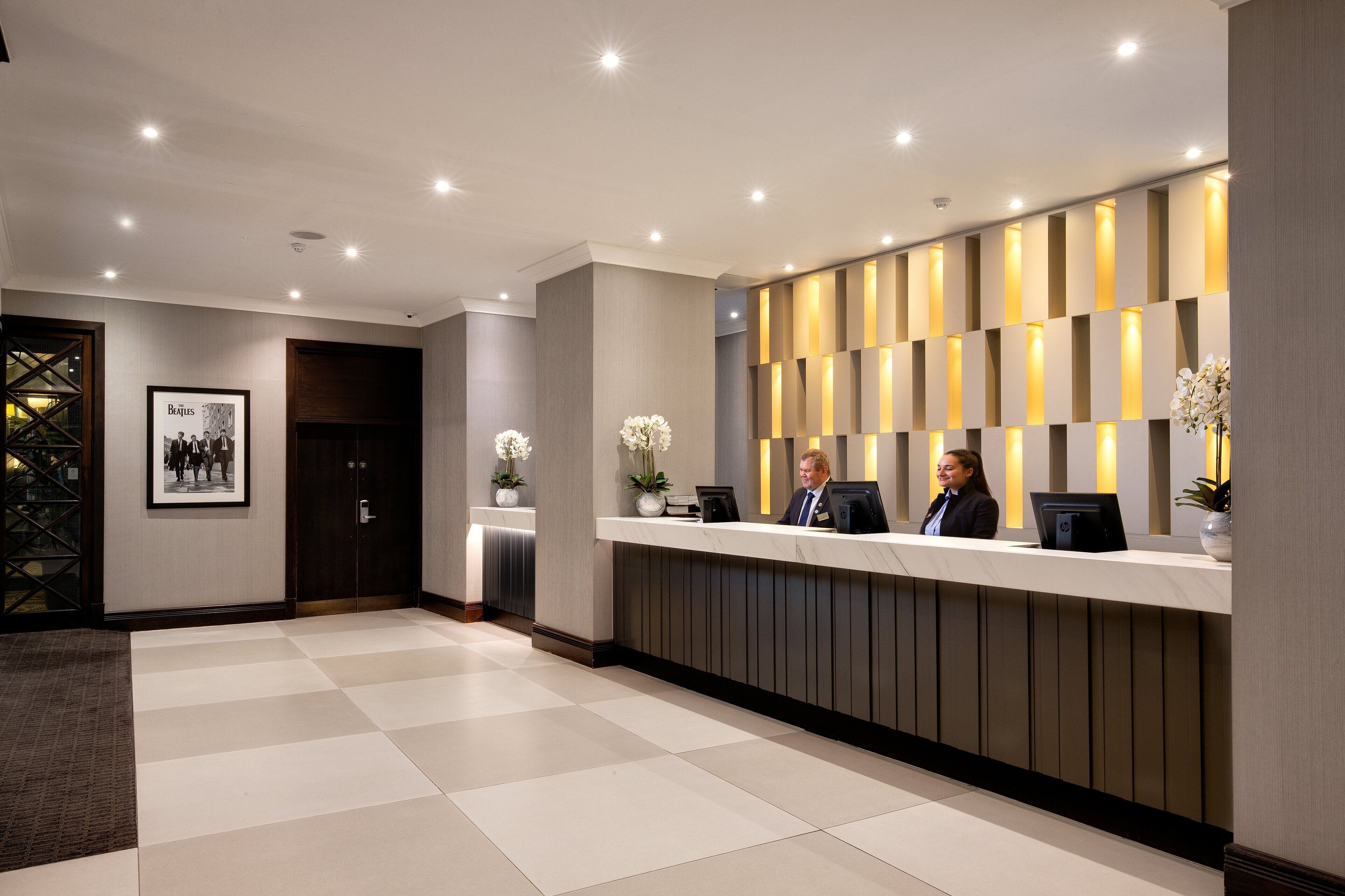 A reception area with a check-in counter, two receptionists, framed photographs on the wall, and potted plants.