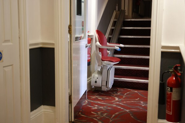 A hotel corridor with a red carpet, a fire extinguisher, and a wheelchair. - OYO Tong Park Hotel (Bradford)