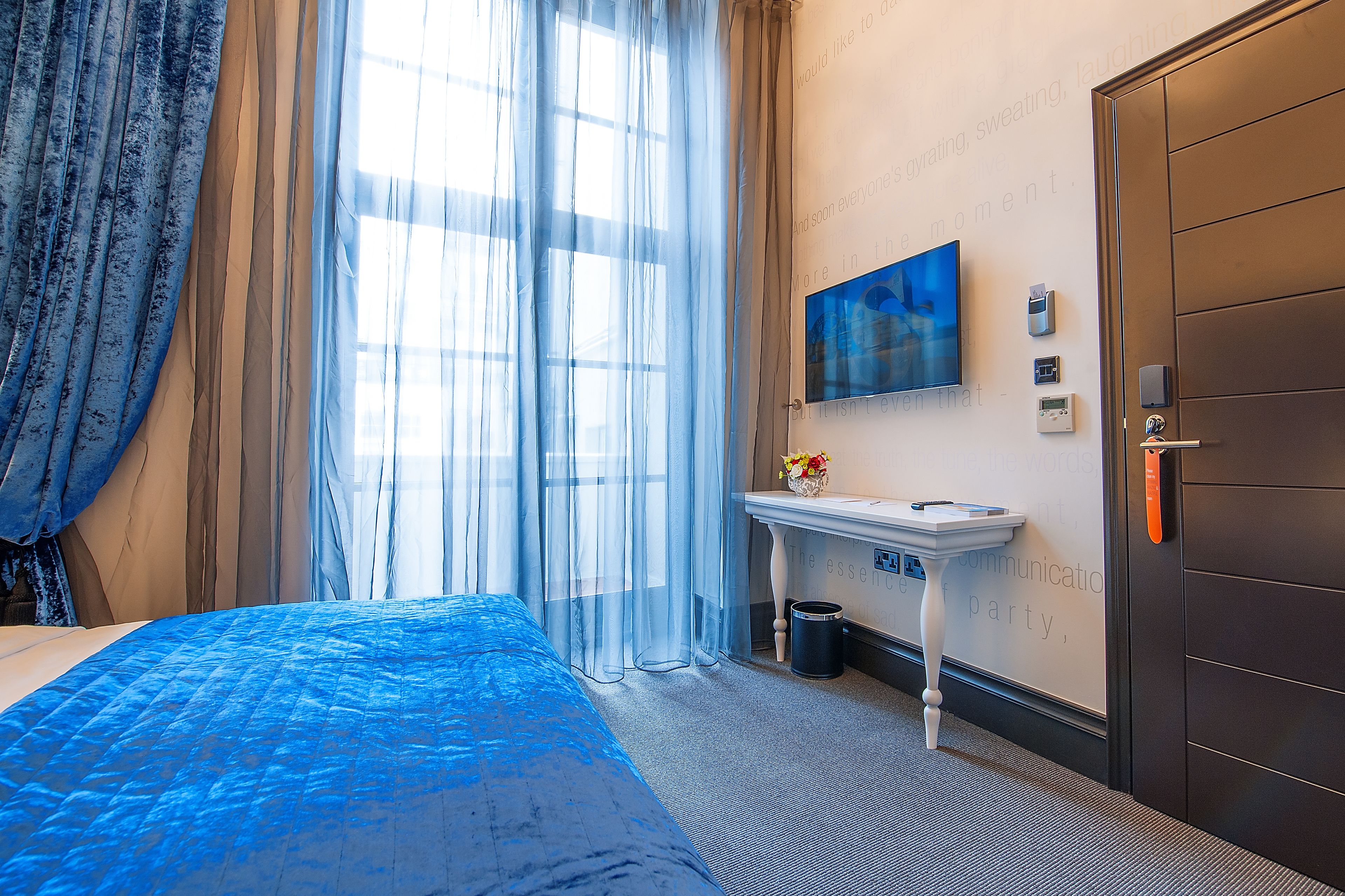 A hotel room with a bed, a small table with a vase, a wall-mounted TV, and a door with a handle.