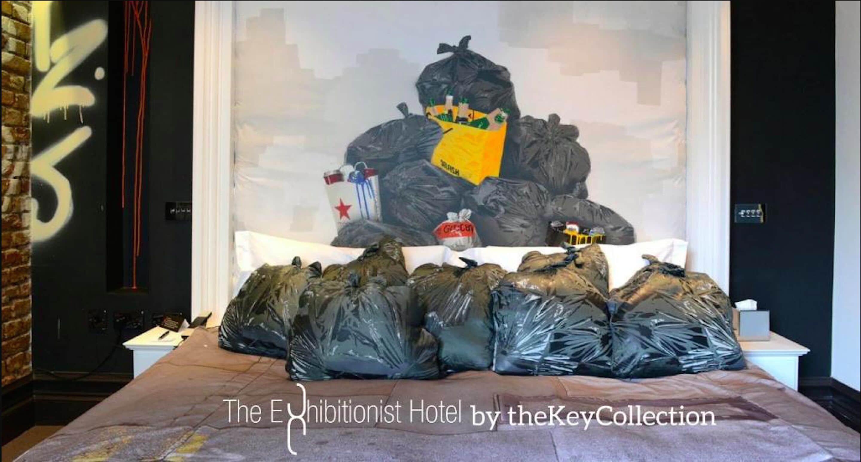 Ein Hotelzimmer mit einem Bett, das mit schwarzen Plastiksäcken abgedeckt ist, einem großen Gemälde an der Wand und einer Backsteinmauer links.
