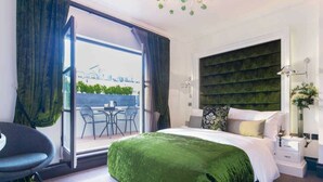A modern bedroom with a large bed, a view of the city through a sliding door, a desk with a chair, and a nightstand with a lamp. - The Exhibitionist Hotel (London)