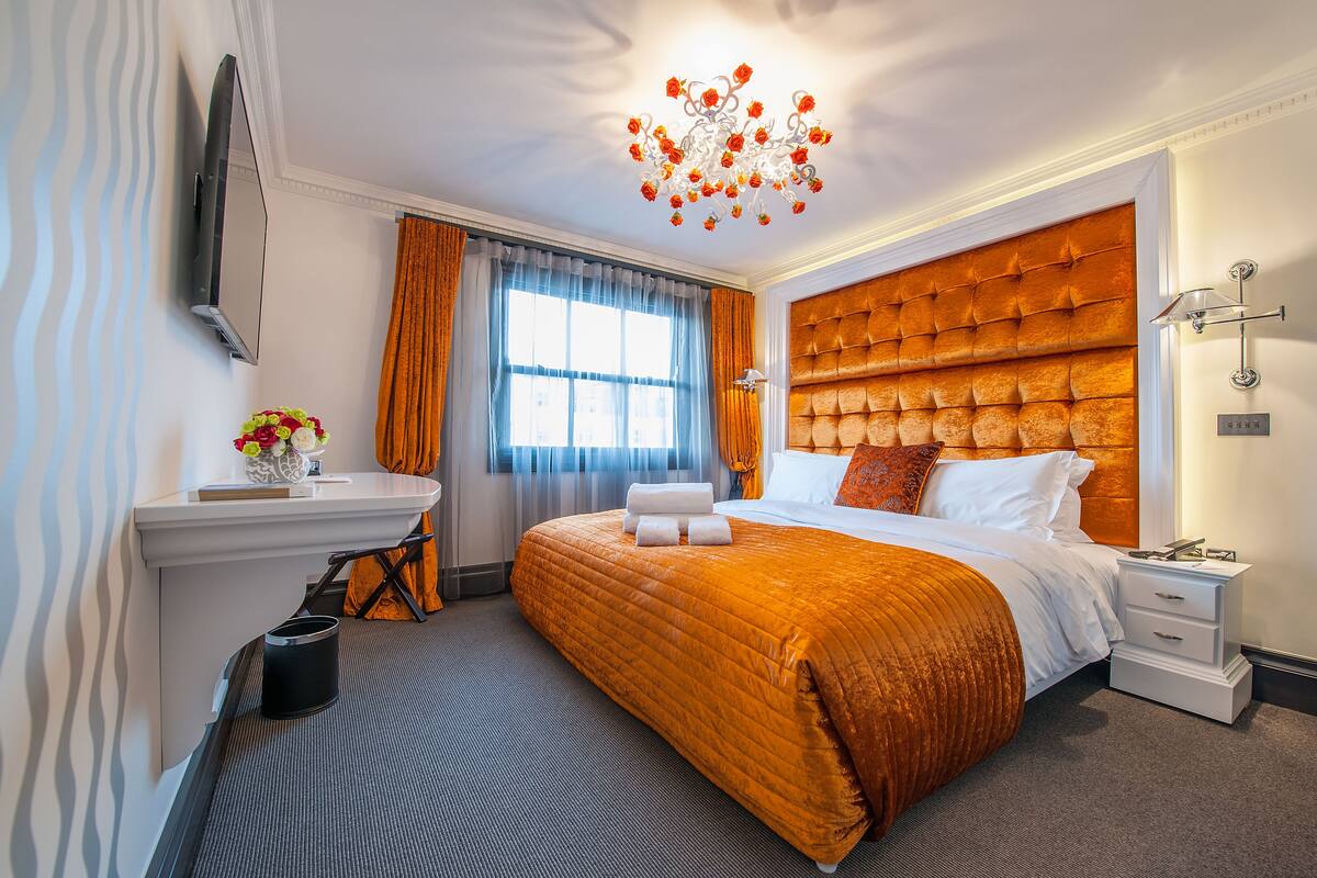 Habitación de hotel con una cama grande, una mesita de noche, un televisor montado en la pared y un ventanal con cortinas.