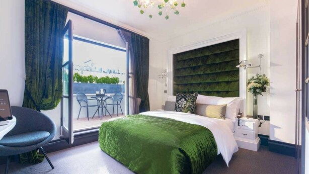 A modern hotel room with a large bed, a desk, and a view of the cityscape through the window. - The Exhibitionist Hotel (London)