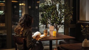 Una persona leyendo un libro en una mesa con una planta, un vaso de jugo de naranja y un tarro de miel.