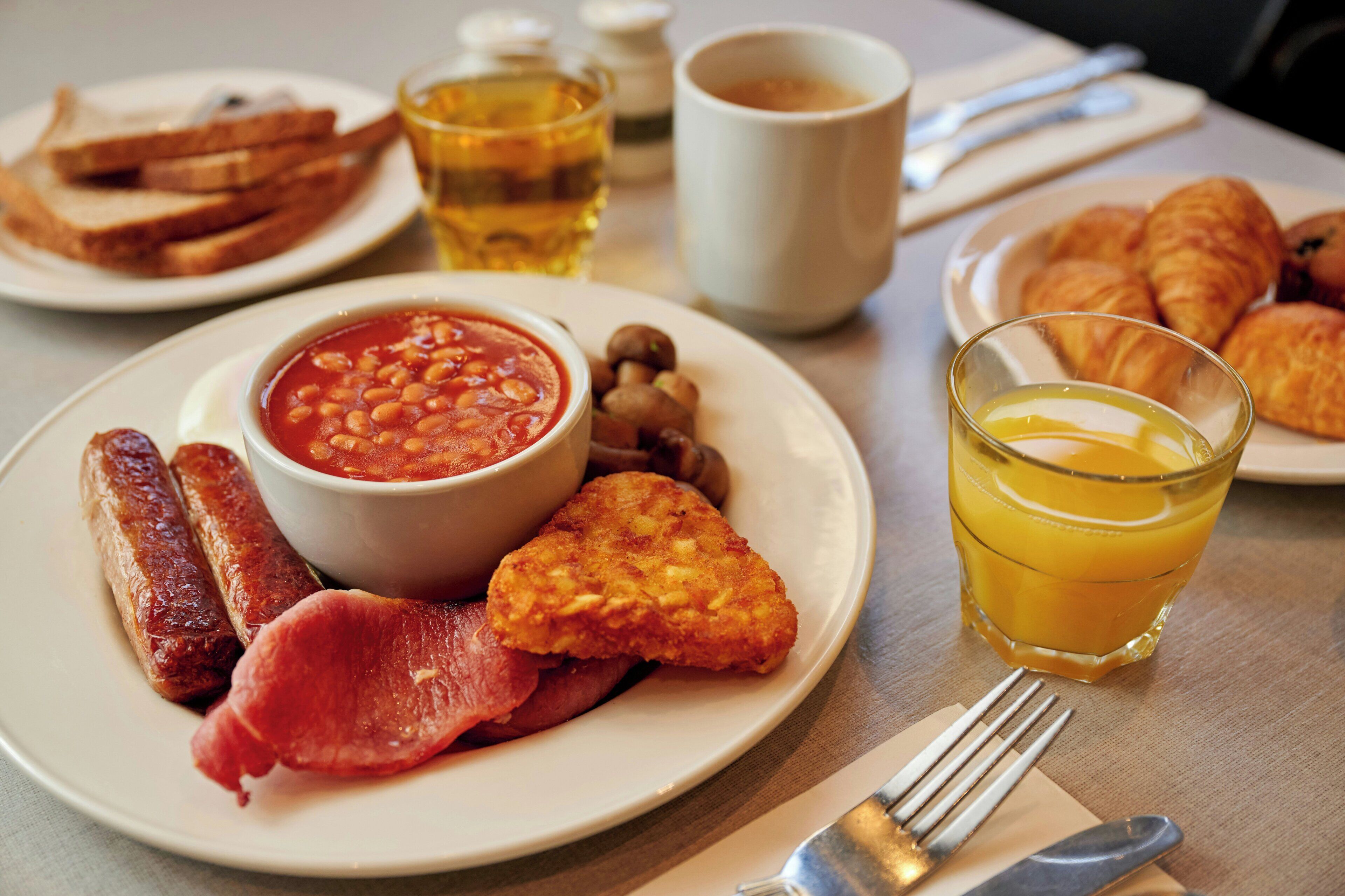 Un plato de desayuno con frijoles, salchichas, tocino, cruasanes, tostadas y un vaso de jugo de naranja.