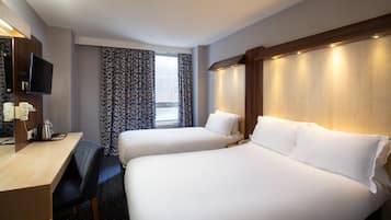Habitación de hotel con dos camas, un escritorio con silla, televisión y ventana con cortinas estampadas.
