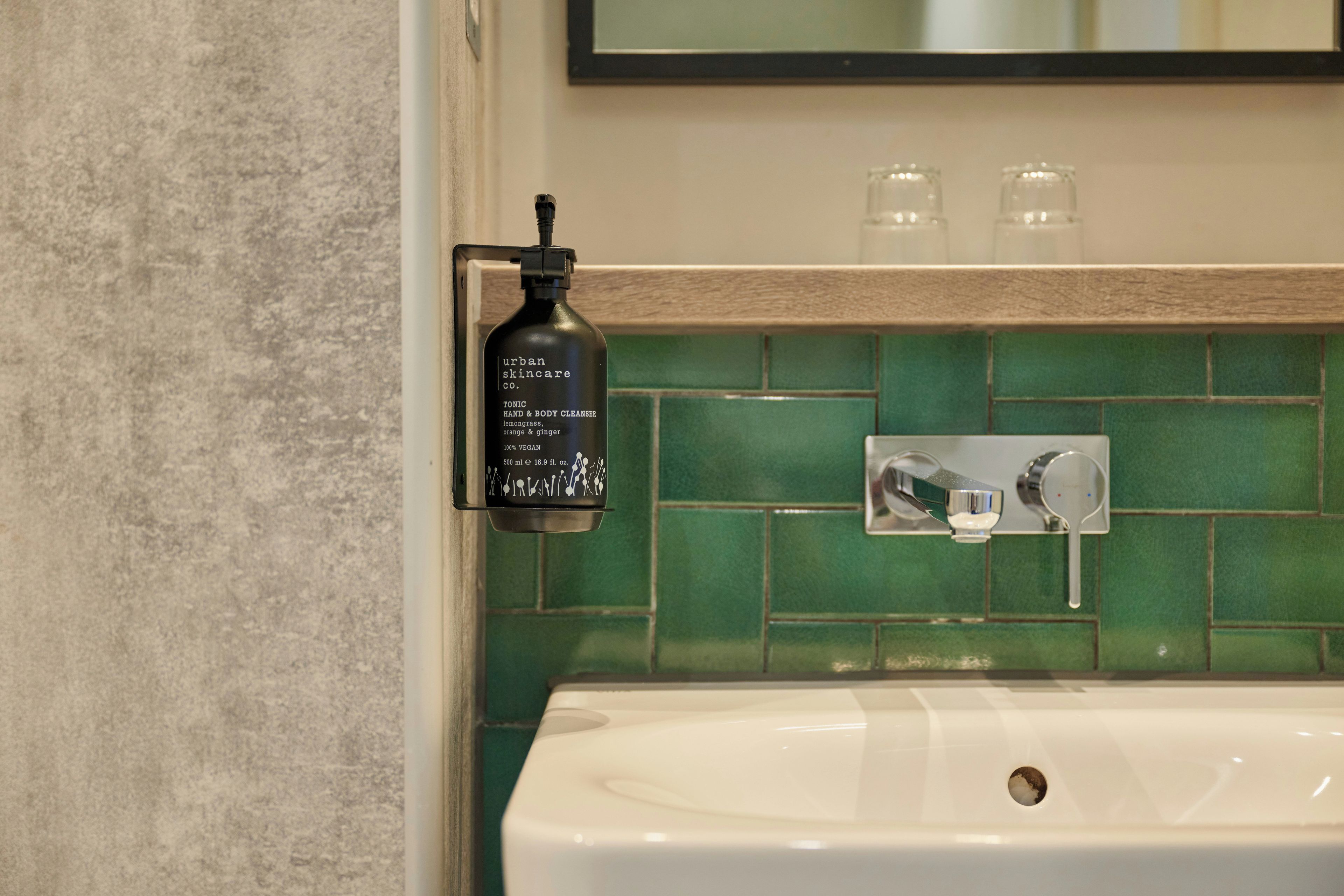 Un baño con un lavamanos blanco, un salpicadero de azulejos verdes y una botella negra de tónico limpiador de manos y cuerpo Urban Skincare.