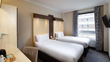 A hotel room with two beds, a desk with a coffee maker, and a view of a building through the window.
