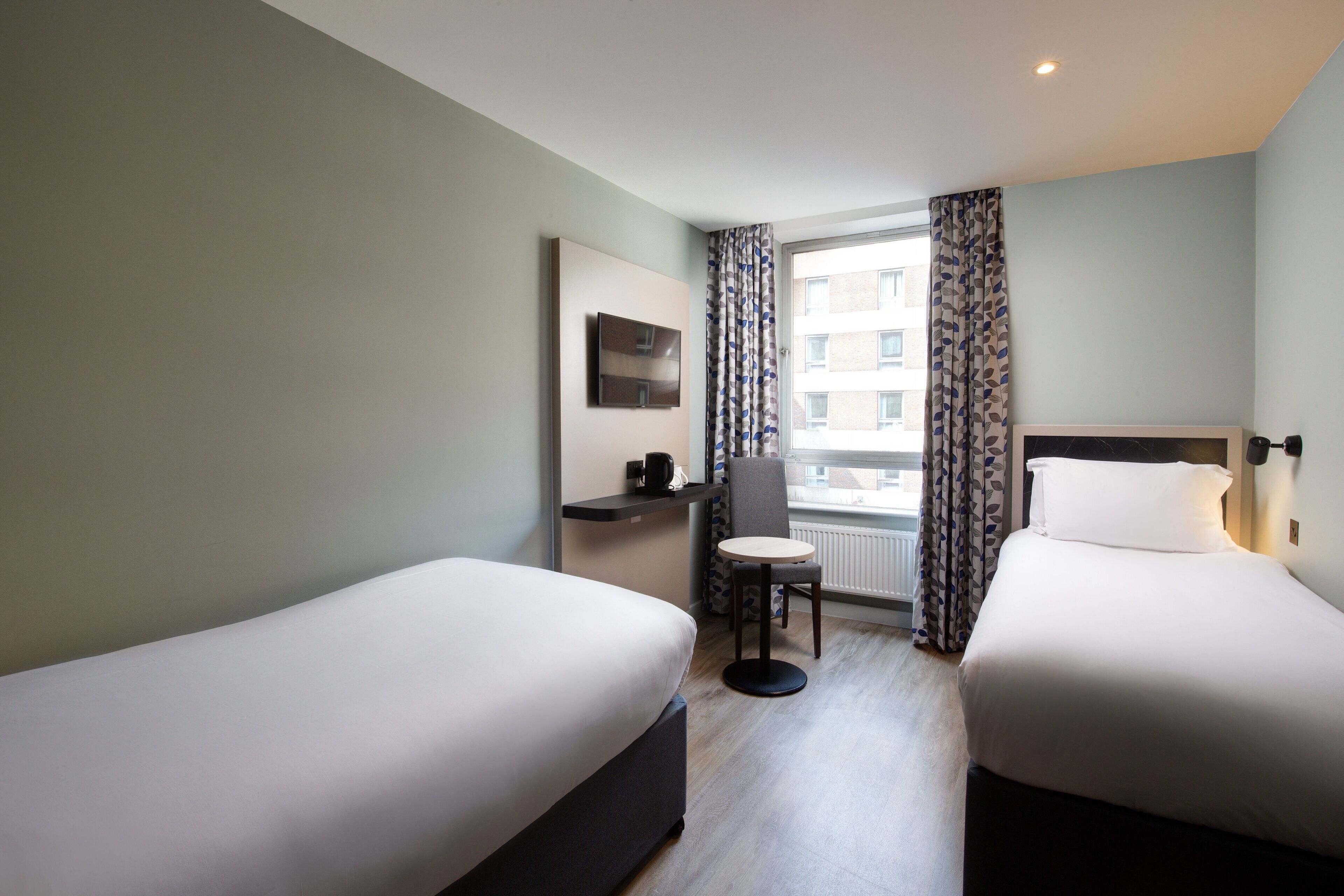 A hotel room with two single beds, a small table, a chair, a TV, and a window with curtains.