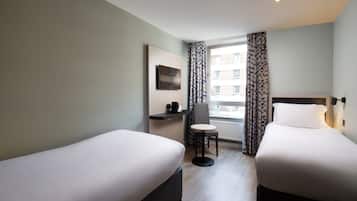 Ein Hotelzimmer mit zwei Einzelbetten, einem kleinen Tisch, einem Sessel, einem Fernseher und einem Fenster mit Vorhängen.