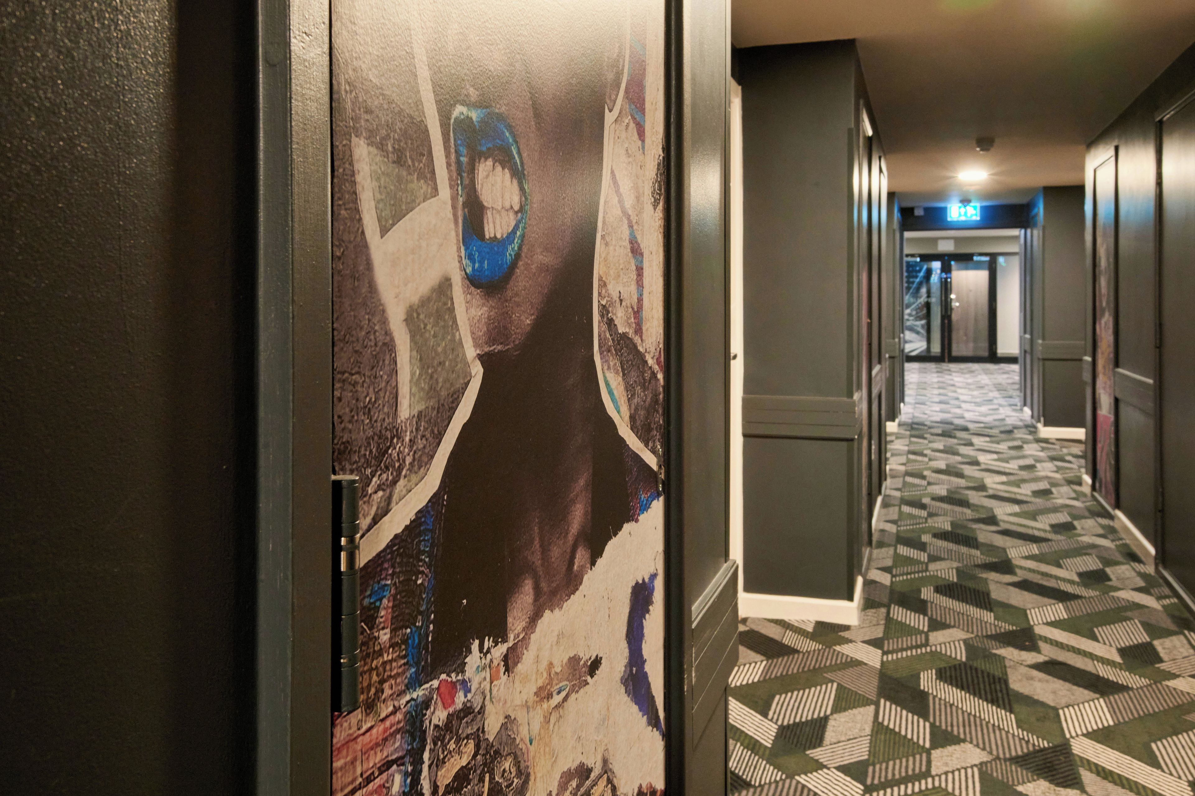 Een hotelgang met een getextureerd tapijt, een muur met een groot muurschilderij, en een deur met een deurklink.